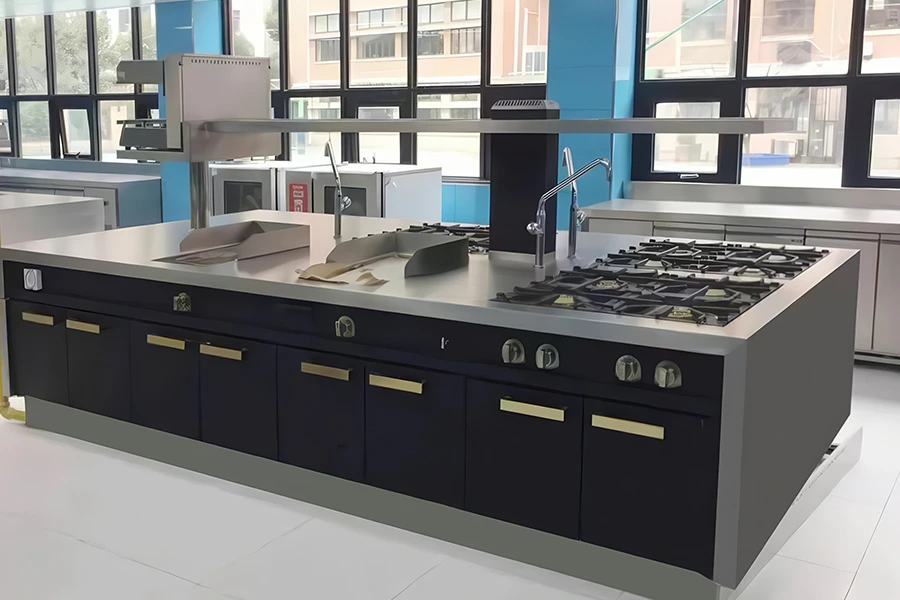 A commercial kitchen with black lower cabinets, stainless steel countertops, and multiple gas burners. Island cooking suites enhance the workspace, while large windows provide natural light.