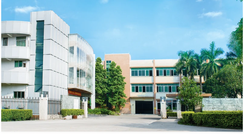 A modern, light-colored building complex with multiple stories stands as a welcoming home, surrounded by greenery and a clear blue sky in the background. There is an open courtyard and a gated entrance in the foreground.