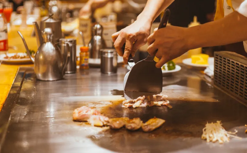 A chef expertly cooks meat on a teppanyaki grill, skillfully wielding metal spatulas to prepare the meal. Various condiments and tools are visible in the background, enhancing the culinary scene.