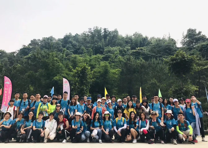 A large team in blue shirts poses for a group photo outdoors, with trees and hills in the background. Some are holding flags, and everyone appears to be smiling, showcasing their spirit of collaboration.