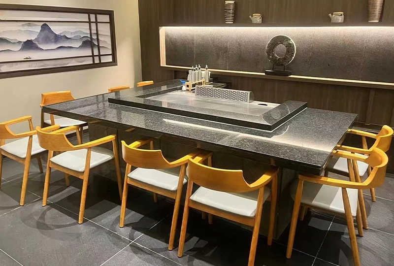 A modern dining area features a large black granite table, perfect for teppanyaki, surrounded by wooden chairs with white cushions. A grill is embedded in the center of the table. Minimalist decor and wall art complete the stylish look.