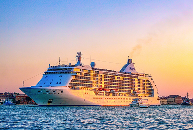 A large cruise ship with a service guarantee sails near a harbor under a colorful sunset sky, with a smaller boat navigating the water in front of it.