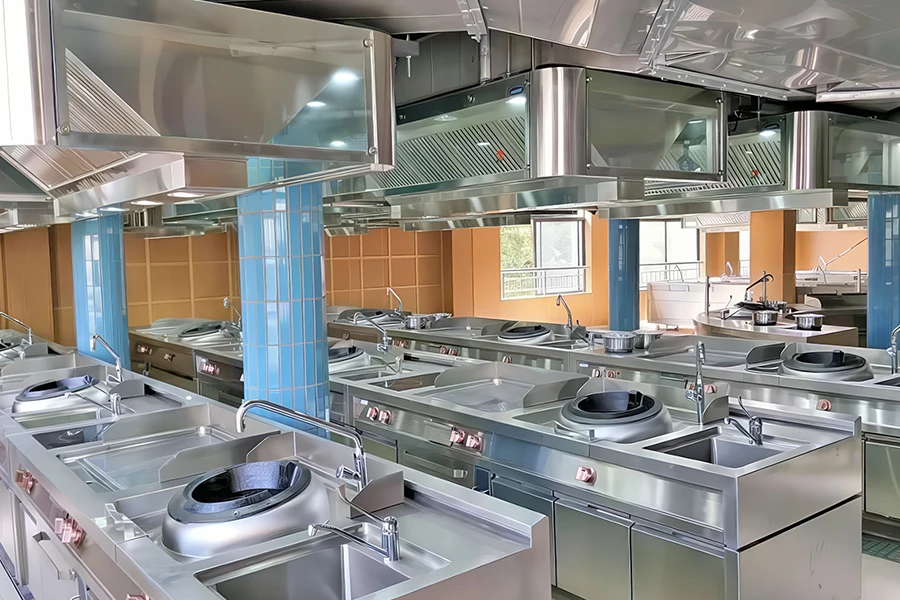 A modern, stainless steel commercial kitchen with multiple workstations, sinks, and cooking ranges arranged in rows, featuring state-of-the-art commercial cooking equipment, overhead ventilation systems, and a light blue tiled column.