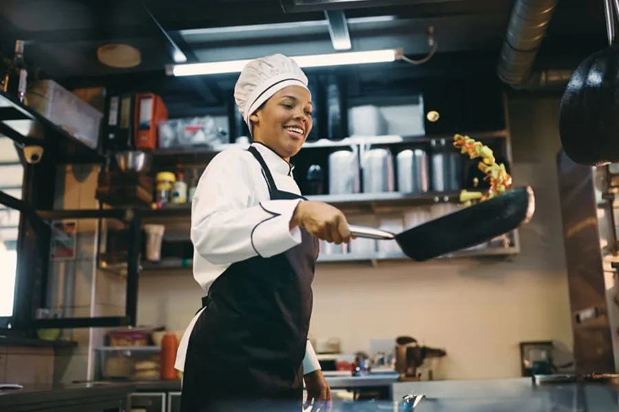 A chef in a white hat and black apron is flipping vegetables in a frying pan while cooking on a commercial induction cooking range.