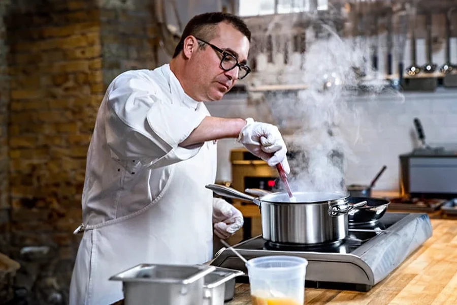 A chef in a white uniform stirs a steaming pot on a stove, the aroma filling the rustic kitchen setting enhanced by the precision of a commercial induction cooking range.