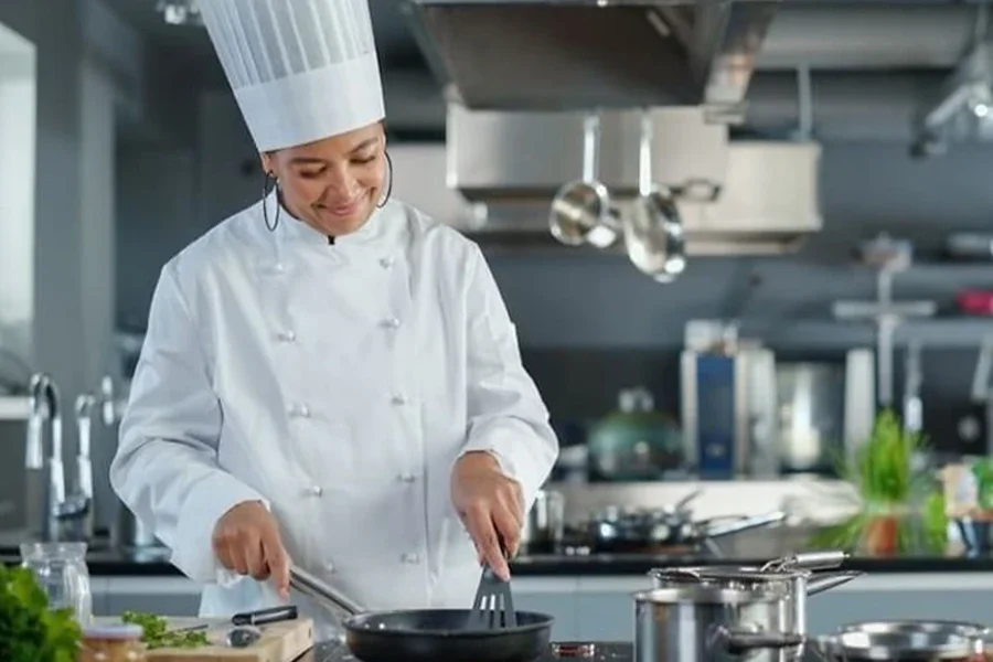 A chef in a white uniform and tall hat cooks in a professional kitchen, smiling and engaging with a pan on the stove. Various kitchen utensils, ingredients, and sleek modular cooking equipment are visible around her.