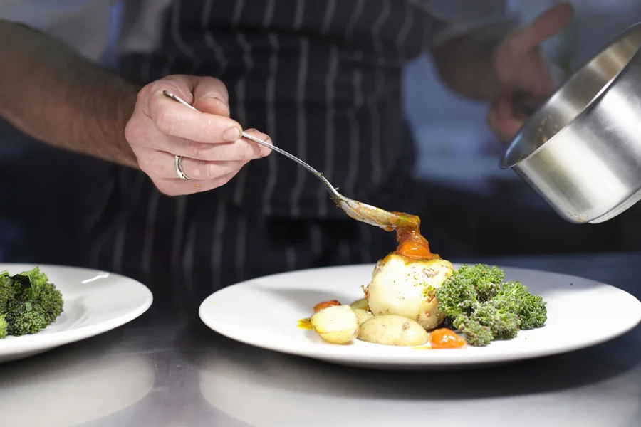 A person in a striped apron, using restaurant cooking equipment, pours sauce over a plated dish with potatoes and broccoli.