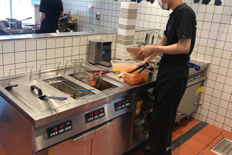 A worker in a kitchen is preparing food next to commercial cooking equipment, including a deep fryer. Stacked food containers and a tiled wall are visible in the background.