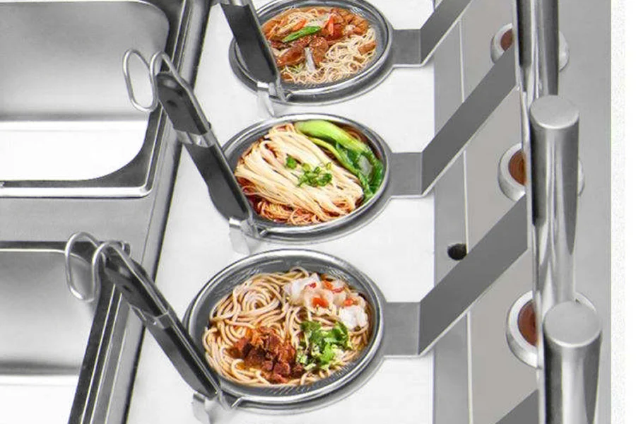 Three bowls of noodles with various toppings are placed in a stainless steel food counter, each with a large spoon inserted. Prepared using our state-of-the-art commercial noodle cooker, these dishes promise both efficiency and taste.