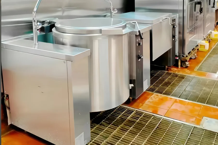 Commercial kitchen with large stainless steel cooking equipment, including a tilt skillet, Tilting Boiling Pan, and industrial stove, on a tiled floor with drains.