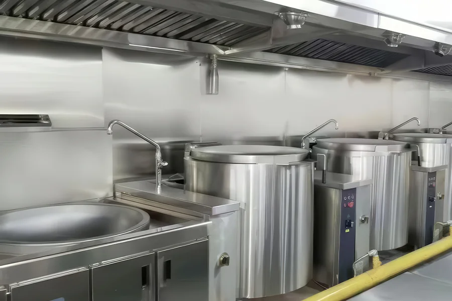 The commercial kitchen is equipped with stainless steel cooking equipment including multiple large cooking pots, a tilting pot, a faucet and an overhead exhaust system.