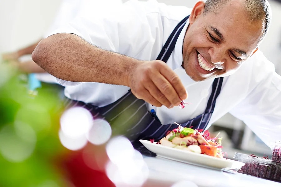 A chef in a white uniform and striped apron smiles while garnishing a plate of food with herbs in a kitchen, standing beside a Commercial Induction Cooker.