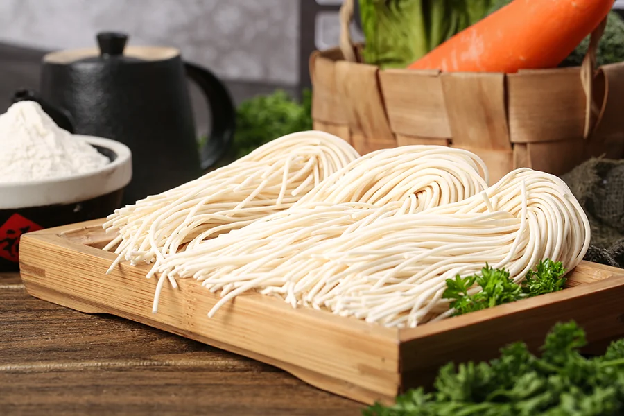 Wooden tray with neatly arranged bundles of uncooked noodles, surrounded by fresh parsley. A basket with vegetables, including a carrot, and a container of flour are also visible in the background, hinting at the perfect setting for using a Commercial Noodle Cooker.