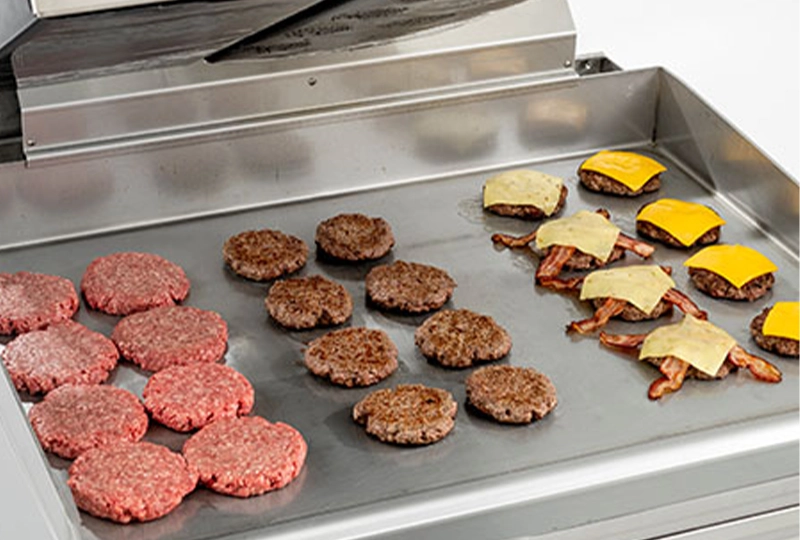 Commercial Clamshell Griddle cooking hamburgers in various stages: raw patties, partially cooked, and fully cooked with melted cheese and bacon, demonstrating efficient batch cooking on a stainless steel surface