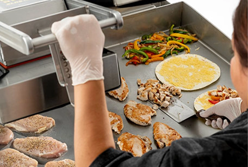 A chef using a commercial clamshell griddle to cook various foods, including seasoned chicken breasts, chopped chicken, sautéed peppers, and tortillas with cheese, highlighting the versatility and efficiency of the equipment for preparing multiple ingredients simultaneously