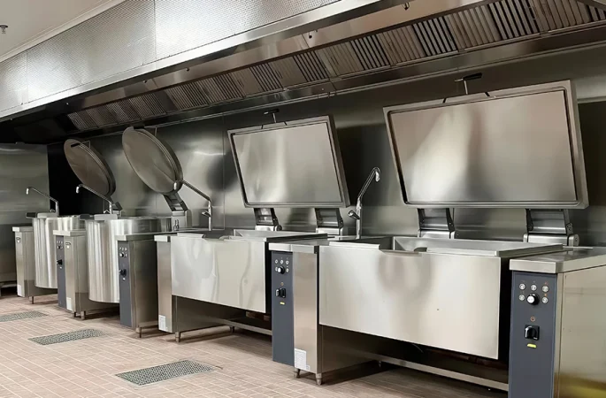 Tilting Bratt Pan cooking equipment in a commercial kitchen, showing a row of modern stainless steel cooking equipment with tilting lids and control panels, suitable for large batch cooking.