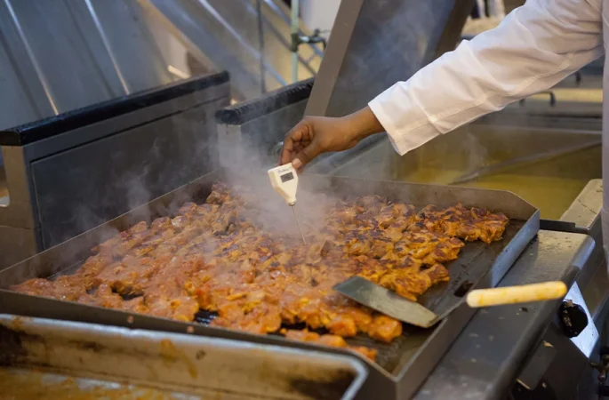 A person in a white coat uses a thermometer to check the temperature of meat cooking on a large commercial griddle, with visible steam rising from the meat.