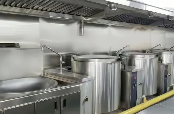 The commercial kitchen is equipped with stainless steel cooking equipment including multiple large cooking pots, a tilting pot, a faucet and an overhead exhaust system.