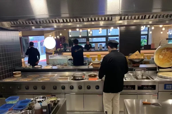 Chefs work in a modern, open kitchen with marbled countertops and stainless steel equipment. One chef tends a large pot on an induction cooker, while others prepare dishes at a rear counter.